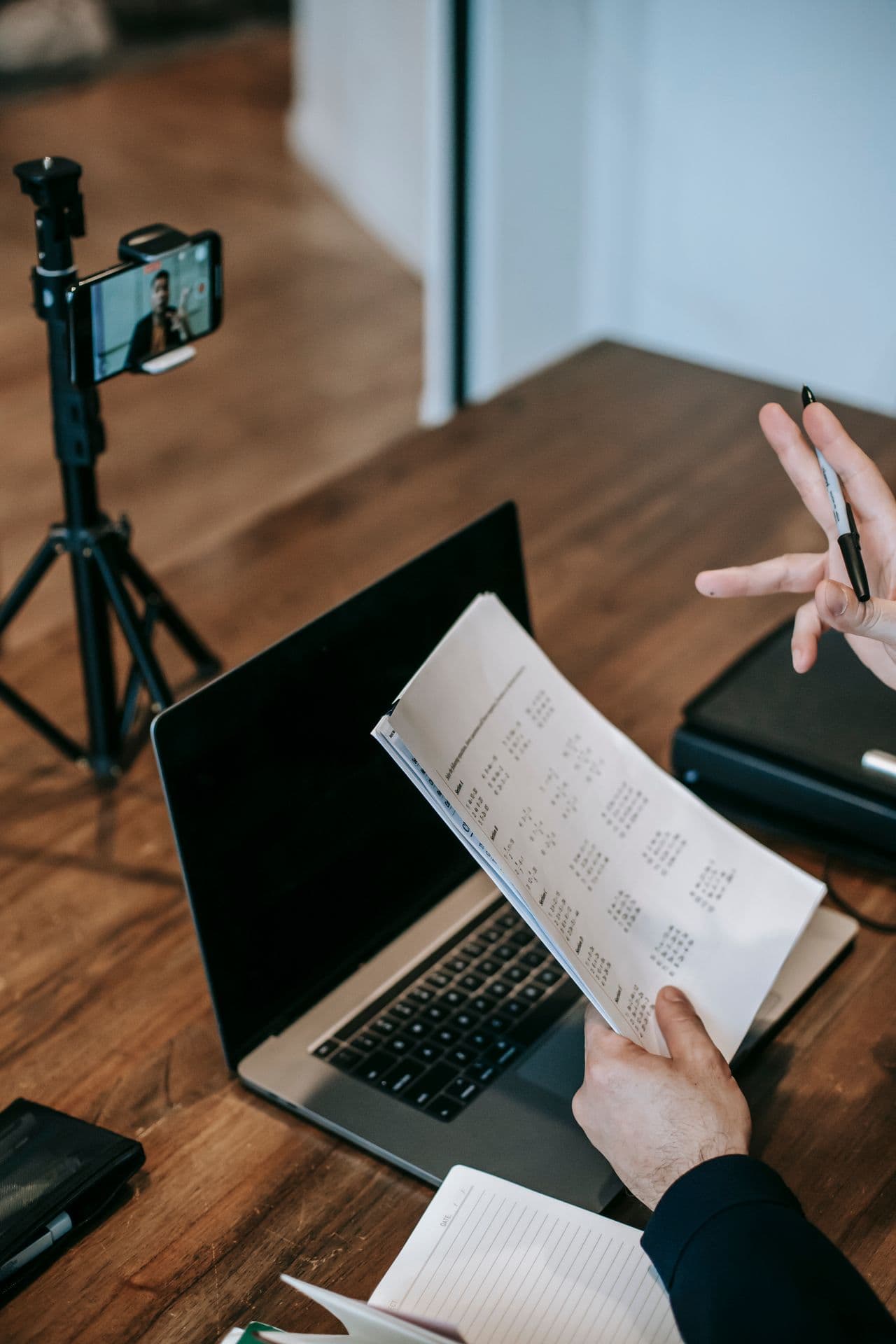 A tutor at a laptop reviewing a printed sheet of a student's marks, with a phone on a tripod set up for the lesson