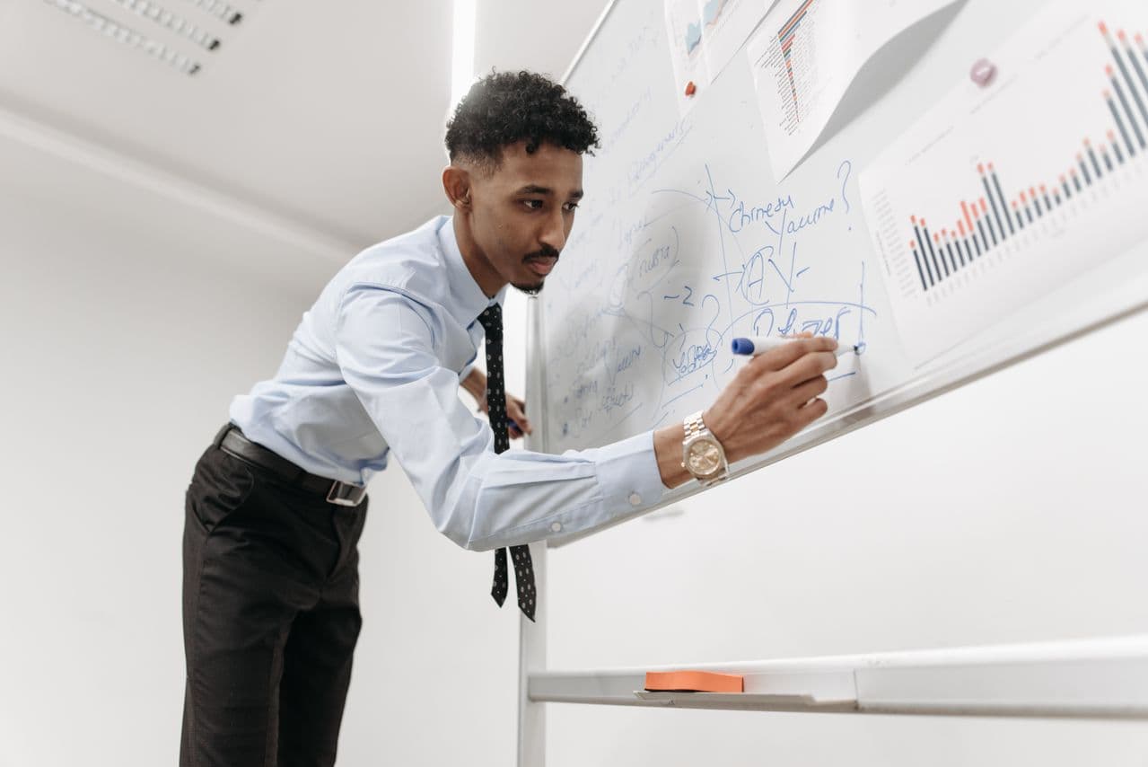A tutor working through a problem in marker on a whiteboard covered in handwritten notes and printed charts